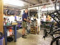 A person adjusting a bicycle in a bike repair shop, surrounded by tools, bicycle parts, and hanging tires. The shop features a workbench with various items and equipment organized around the space.