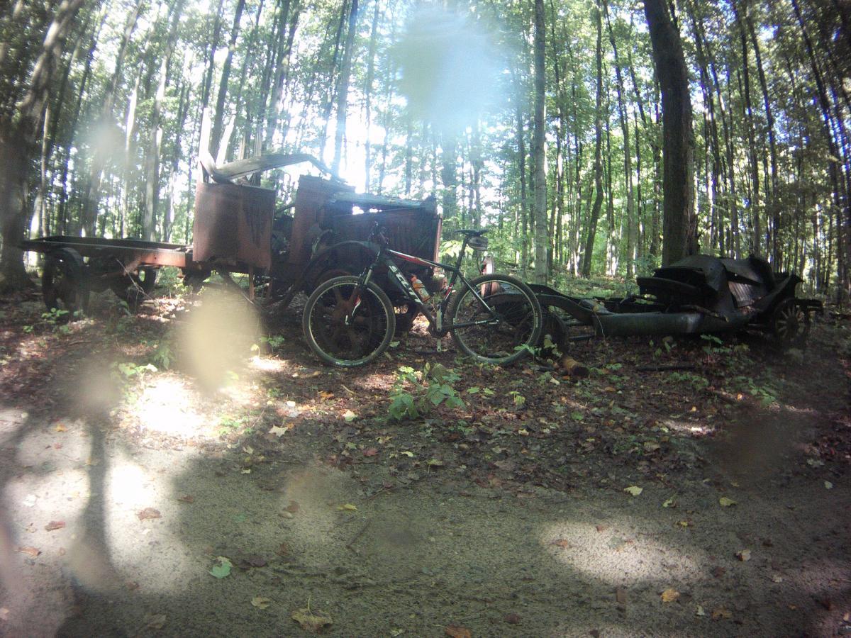 A rusty, vintage truck and a partially dismantled vehicle are parked in a wooded area, surrounded by tall trees. There is a bicycle leaning against the truck, and the forest floor is covered with leaves and small plants. Natural sunlight filters through the trees, creating a dappled effect on the ground. Louis M. Groen Nature Preseve mountain bike trail.