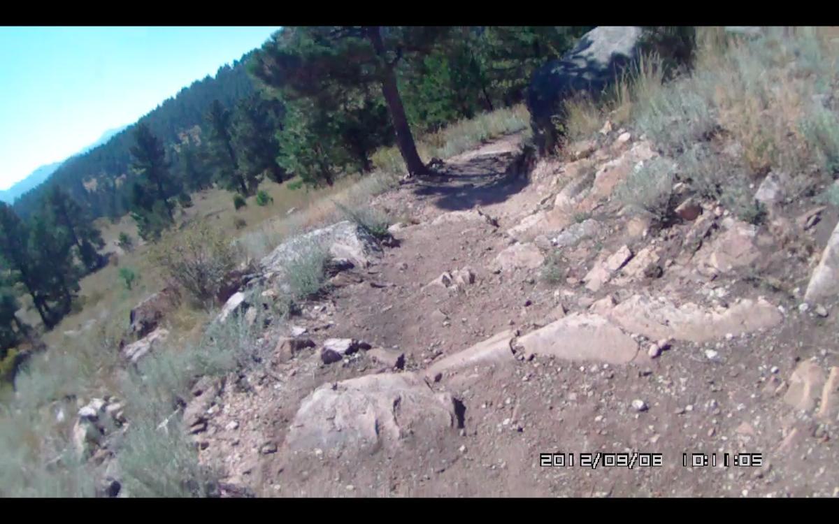 A dirt trail winding through a mountainous landscape, surrounded by trees and rocky outcrops, under a clear blue sky. The scene captures the natural beauty of an outdoor hiking path. Elk Meadow mountain bike trail.