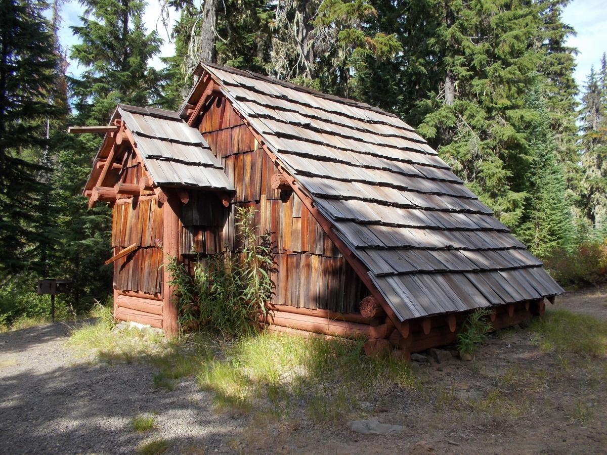 A rustic wooden cabin with a sloped roof, surrounded by tall evergreen trees. The cabin features weathered wood siding and a small protruding structure on one side. The setting is a natural forested area, with a gravel path leading up to the cabin. Gold Lake To Bobby Lake mountain bike trail.