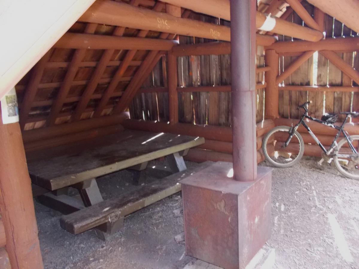 An interior view of a rustic shelter made from wooden logs, featuring a wooden picnic table, a metal stove, and a mountain bike leaning against the wall. The structure has a sloped roof and open walls, allowing natural light to filter in. Gold Lake To Bobby Lake mountain bike trail.