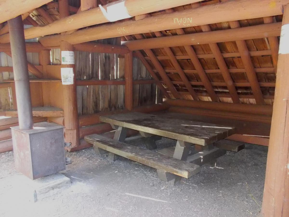 A rustic wooden shelter featuring a picnic table made of dark wood, with a metal stove in one corner. The interior walls are lined with wooden beams, and there are some signs attached to the walls. Sunlight filters in, illuminating the simple, natural surroundings. Gold Lake To Bobby Lake mountain bike trail.