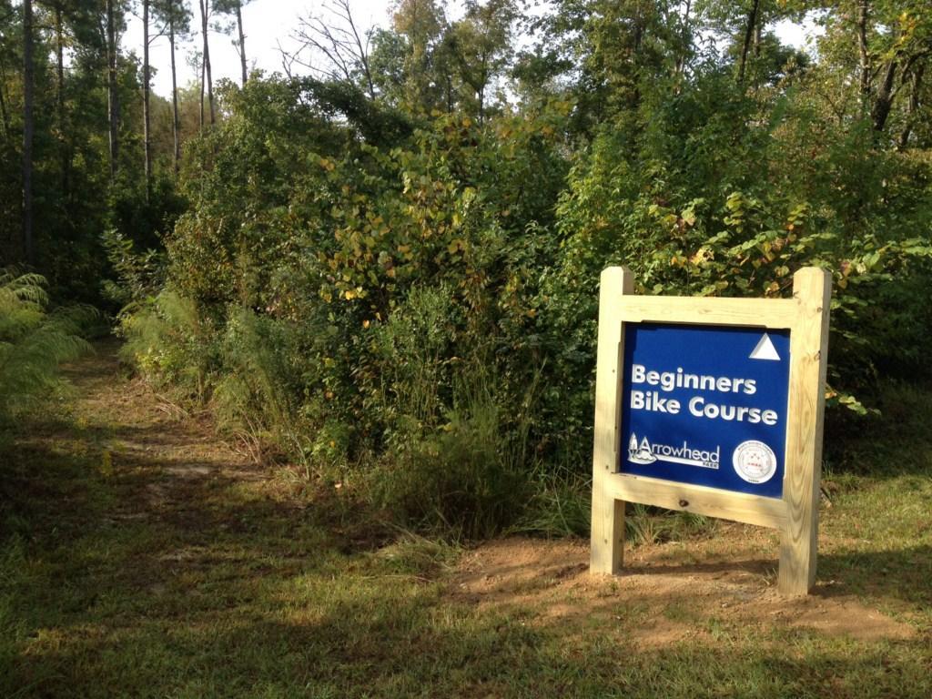 A sign for a beginners bike course is positioned at the entrance of a wooded area, surrounded by lush greenery and trees. The trail path leads into a natural setting, inviting cyclists to explore. Arrowhead Park mountain bike trail.