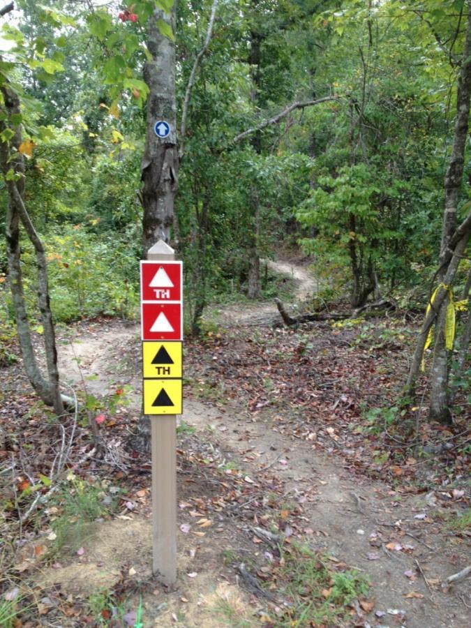 Signpost at a trailhead in a wooded area, displaying directional markers and trail difficulty indicators. Red and yellow triangular signs labeled "TH" guide hikers towards the paths diverging into the forest. The surrounding trees feature green foliage, and a dirt trail is visible winding through the underbrush. Arrowhead Park mountain bike trail.
