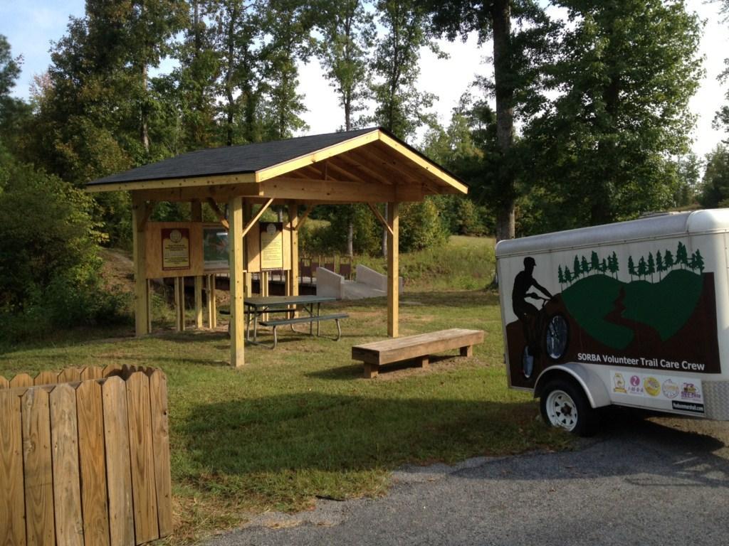 A wooden shelter with a picnic table underneath, located in a grassy area surrounded by trees. Next to the shelter, there is a small trailer marked with "SORBA Volunteer Trail Care Crew" and an illustration of a cyclist. A wooden fence is visible in the foreground. Arrowhead Park mountain bike trail.