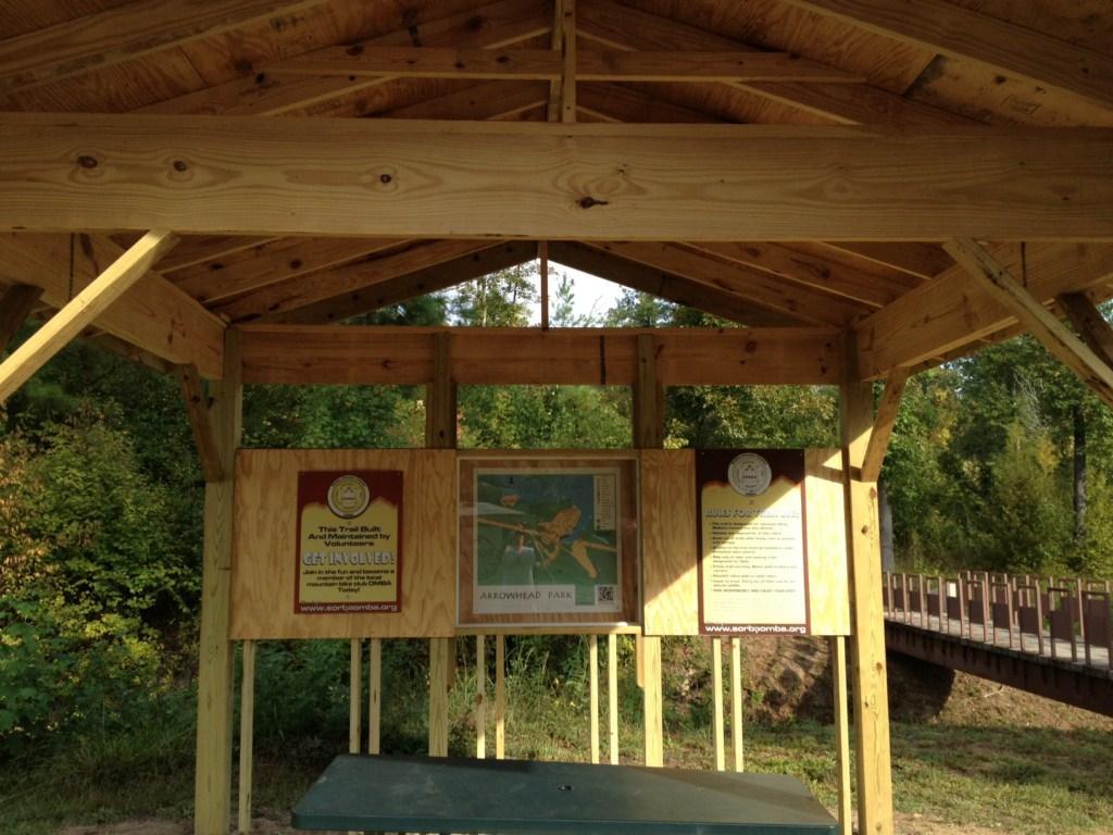 Wooden shelter displaying informational signs about Arrowhead Park, featuring a map and community engagement details, with a bridge visible in the background and lush greenery surrounding the area. Arrowhead Park mountain bike trail.