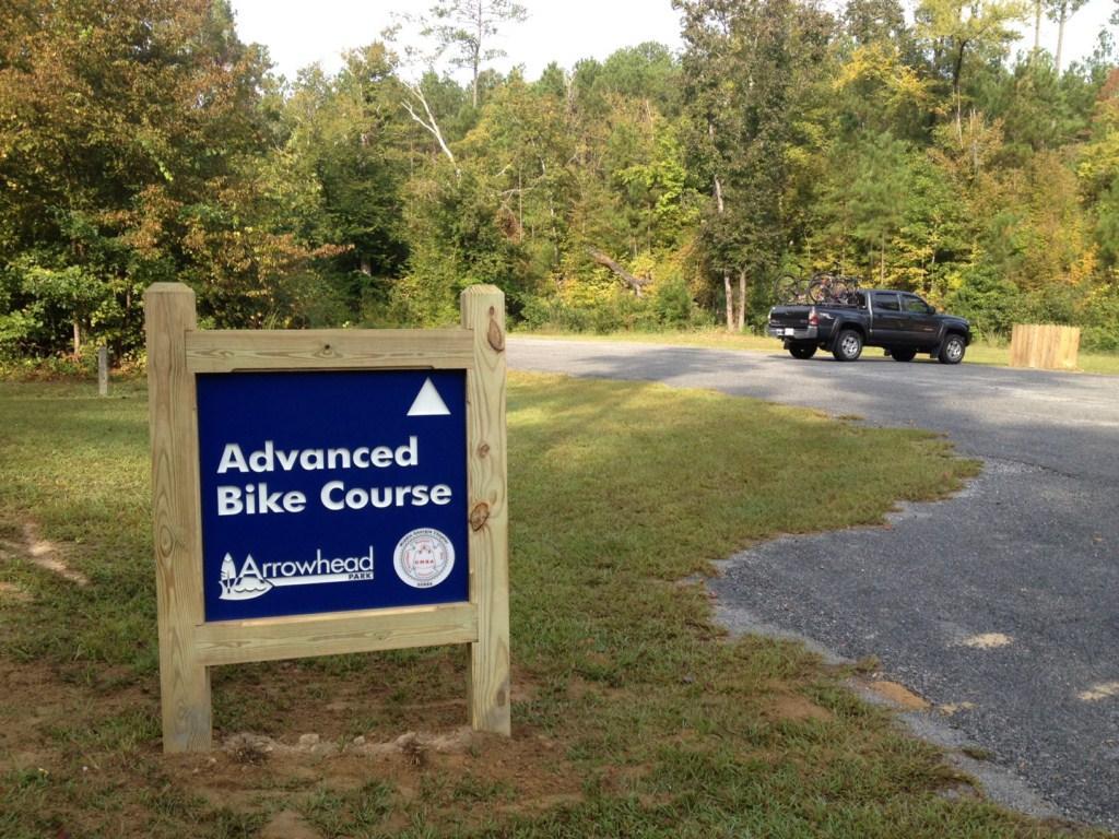 A sign that reads "Advanced Bike Course" stands at the entrance of a wooded area, with a gravel path leading away from it. A black pickup truck with bicycles on the back is parked nearby, surrounded by green grass and trees in the background. Arrowhead Park mountain bike trail.