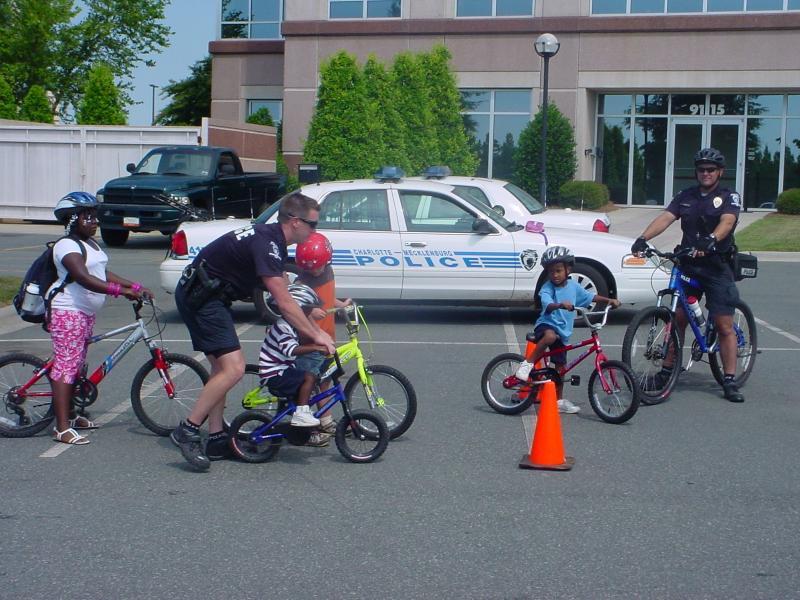 A group of children on bicycles interacts with police officers in a parking lot. Two officers are present, one assisting a child with a red helmet while another officer stands by. The setting includes a police car in the background and trees lining the area. A traffic cone is positioned on the ground among the riders.
