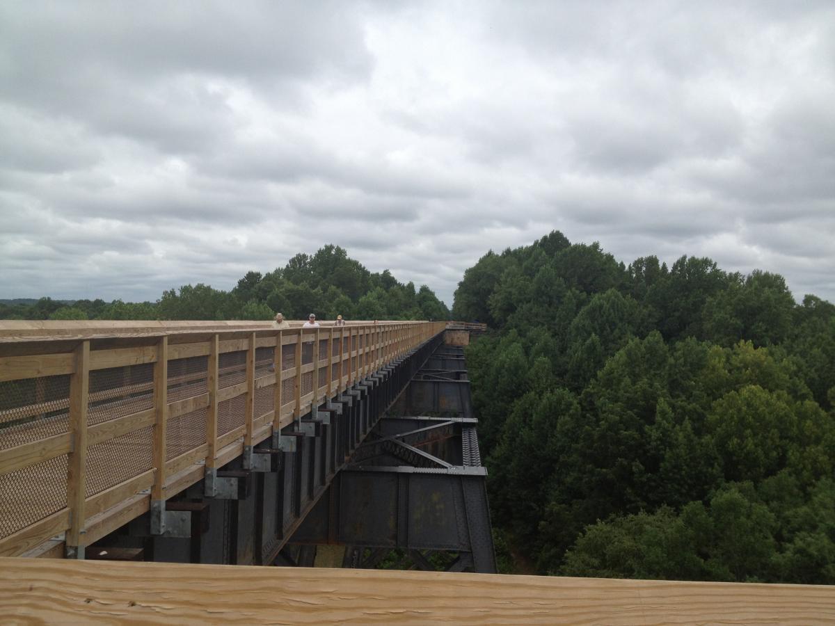 A long wooden pedestrian bridge extends over a lush green landscape, with patches of trees visible on either side. The sky is overcast, featuring gray clouds. A few people can be seen walking along the bridge, enjoying the view of the surrounding scenery. The structure showcases a blend of wood and metal elements. High Bridge Trail mountain bike trail.
