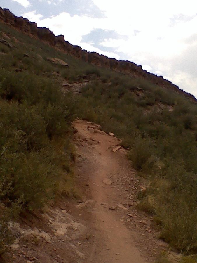 A hiking trail winding uphill through grassy terrain, with rocky outcrops on one side and a cloudy sky above. Blue Sky mountain bike trail.