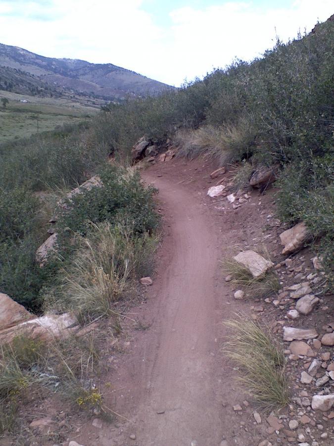 A winding dirt trail framed by shrubs and rocks, leading through a mountainous landscape under a partly cloudy sky. Blue Sky mountain bike trail.
