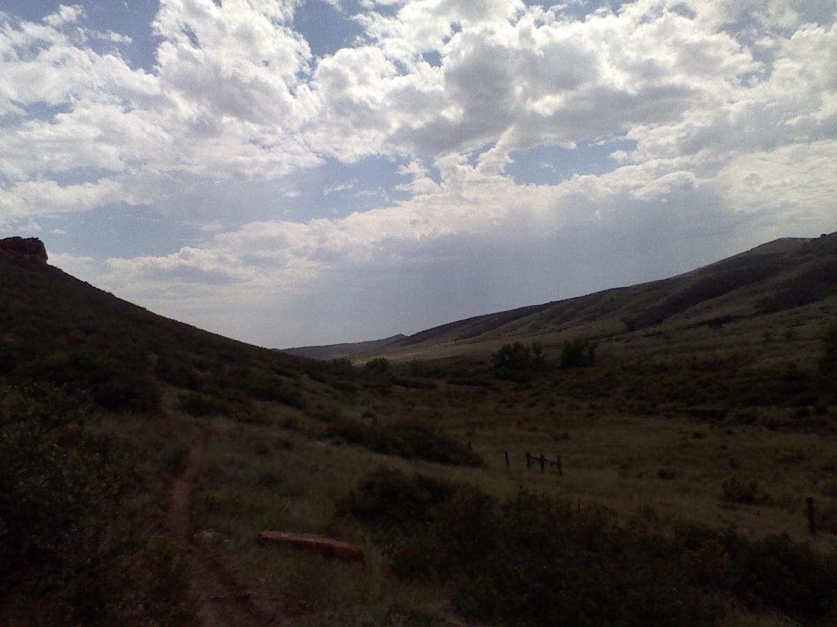 A panoramic view of rolling hills under a partly cloudy sky, with patches of sunlight illuminating the grassy landscape. A dirt path winds through the foreground, leading deeper into the natural scenery. Fences and low shrubbery can be seen in the distance, enhancing the tranquil outdoor atmosphere. Blue Sky mountain bike trail.