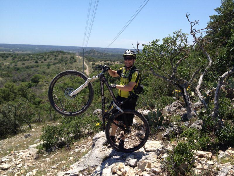 A mountain biker wearing a helmet and a bright yellow and black jersey stands on a rocky outcrop, lifting one wheel of his bike off the ground with a backdrop of a green landscape and clear blue sky. Power lines stretch across the horizon. Flat Rock Ranch mountain bike trail.
