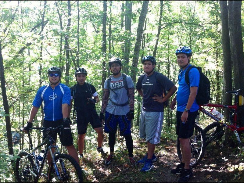 A group of five mountain bikers standing on a wooded trail, wearing helmets and cycling gear. The background features dense green trees and sunlight filtering through the leaves. Bicycles are positioned nearby, and one biker is wearing a race number. Cemetery Loop mountain bike trail.
