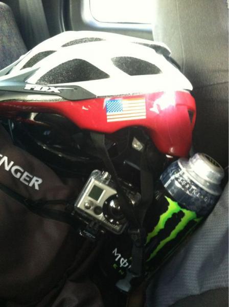 Red bicycle helmet with an American flag sticker, a camera mounted on the side, and a can of Monster energy drink, resting against a backpack in a vehicle. Amelia Earhart Park mountain bike trail.