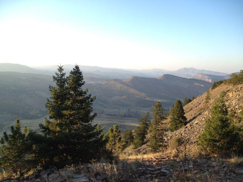 A scenic landscape view featuring rolling mountains under a clear blue sky, with a foreground of evergreen trees and rocky terrain. The sun casts a soft light, highlighting the contours of the hills and valleys below. Heil Valley Ranch mountain bike trail.