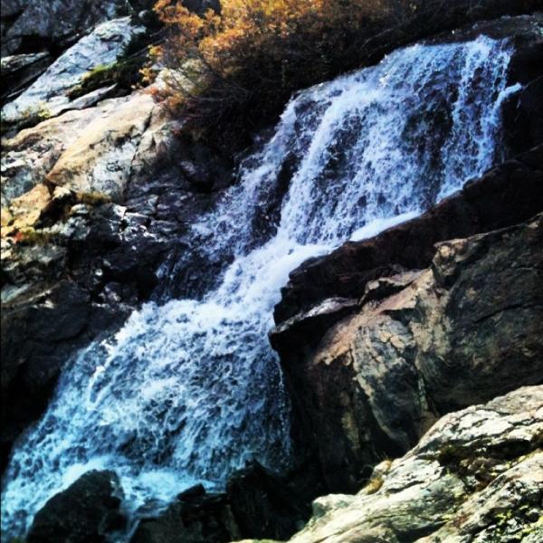 Alt text: A cascading waterfall flows over rugged rocks, surrounded by sparse vegetation. The water is clear and white against the earthy tones of the stones. Mccullough Gulch mountain bike trail.