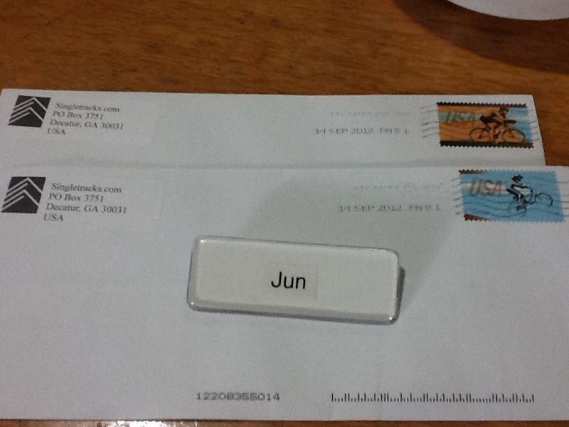 Two envelopes from Singletracks.com with a return address in Decatur, Georgia. Each envelope has a date stamp of September 14, 2012, and features different bicycle-themed stamps. A label with the name "Jun" is placed in front of the envelopes. The background shows a wooden surface. Cemetery Loop mountain bike trail.