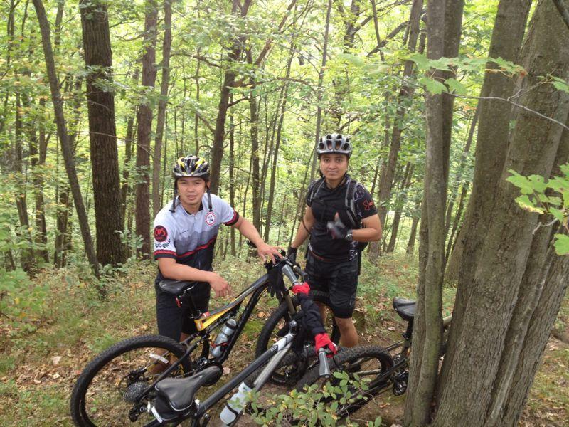 Two mountain bikers pause on a forest trail, surrounded by lush green trees. They are standing next to their bikes, one wearing a helmet and a jersey with a logo, and the other in a black outfit with a helmet. Both are smiling and appear to be enjoying their outdoor adventure. Cemetery Loop mountain bike trail.