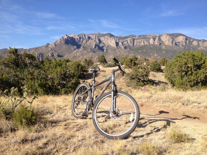 A mountain bike parked on a dirt trail in a grassy area, with mountains in the background under a clear blue sky. The landscape features sparse vegetation and large rocky formations. Elena Gallegos Open Space / North Foothills mountain bike trail.
