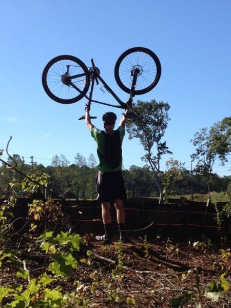 A person wearing a green shirt and black shorts raises a bicycle above their head against a clear blue sky. The background features trees and a wooden fence, suggesting an outdoor setting. Taylor Randahl Memorial Mountain Bike Trails At Olde Rope Mill Park mountain bike trail.
