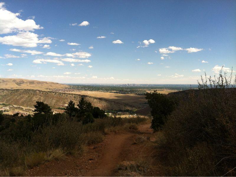 A scenic view from a mountain trail overlooking a vast valley, with a blue sky scattered with clouds. The landscape features rolling hills, patches of greenery, and a distant urban skyline visible in the horizon. A dirt path winds through the foreground. Mount Falcon Park mountain bike trail.