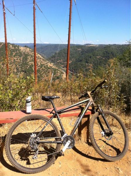 A mountain bike resting on a wooden bench overlooking a scenic valley with green hills, under a clear blue sky. Power lines are visible in the background. A water bottle is placed on the bench beside the bike. Stagecoach / Flood / Manzanita mountain bike trail.