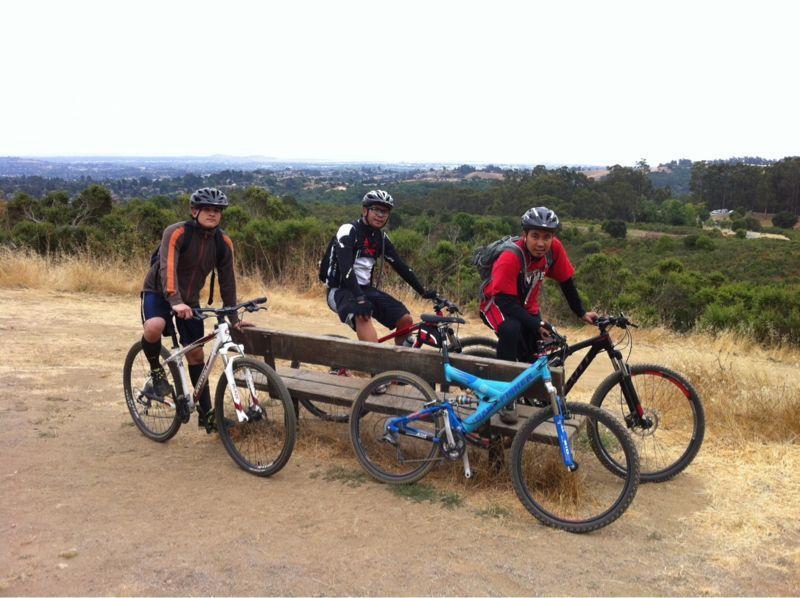 Three mountain bikers in helmets pose with their bikes on a dirt path overlooking a scenic landscape. They are sitting on a wooden bench amid grass and shrubs, with rolling hills visible in the background under a cloudy sky. Anthony Chabot Regional Park mountain bike trail.