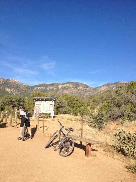 A scenic view of a mountain biking trailhead, featuring a person standing near a sign and a bench, with two bicycles resting against the bench. In the background, there are lush green trees and rugged mountains under a clear blue sky. Elena Gallegos Open Space / North Foothills mountain bike trail.