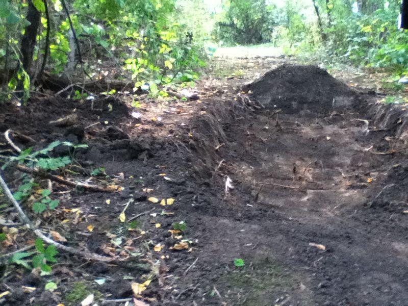 A dirt trail surrounded by greenery, featuring a freshly dug trench on the left side and a mound of earth nearby. The path appears to be in a wooded area with scattered leaves and plants. Hidden heaven mountain bike trail.