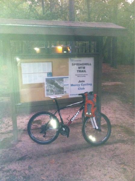 A mountain bike is parked in front of a trailhead sign for the Springhill MTB Trail. The sign includes information about the trail and an invitation to join the Mercy Cycling Club. The setting is wooded, with a slightly foggy atmosphere indicating early morning or late afternoon. Springhill Park mountain bike trail.