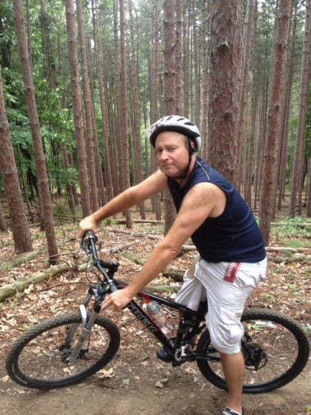 A man wearing a helmet and a sleeveless shirt is sitting on a mountain bike, surrounded by tall trees in a forested area. He appears to be smiling and relaxed while enjoying a moment outdoors. North Park mountain bike trail.