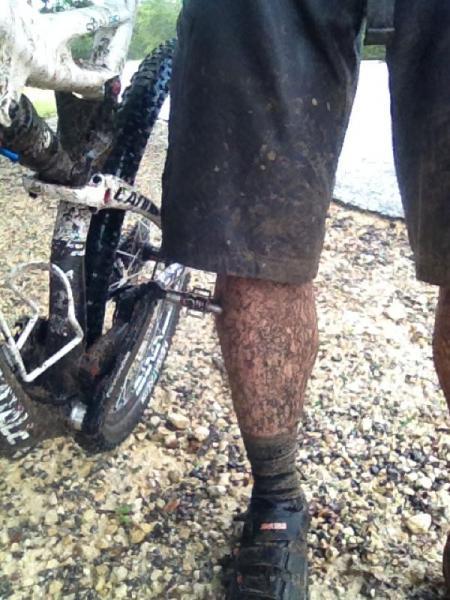 A close-up view of a person's muddy legs and bike, showing dirt caked on shorts and socks after riding in wet, muddy conditions. The image focuses on the bike's rear wheel and the rider's lower body, highlighting the outdoor cycling experience. Madrone Trail mountain bike trail.