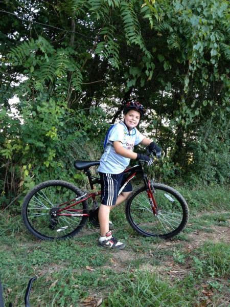 A young child wearing a helmet and casual clothing smiles while posing next to a mountain bike on a grassy path surrounded by greenery. Blue Marsh mountain bike trail.