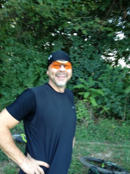 A smiling man wearing a black shirt and a black beanie, with orange-tinted sunglasses, stands in front of lush green foliage. He appears to be enjoying outdoor activity, possibly cycling. Blue Marsh mountain bike trail.