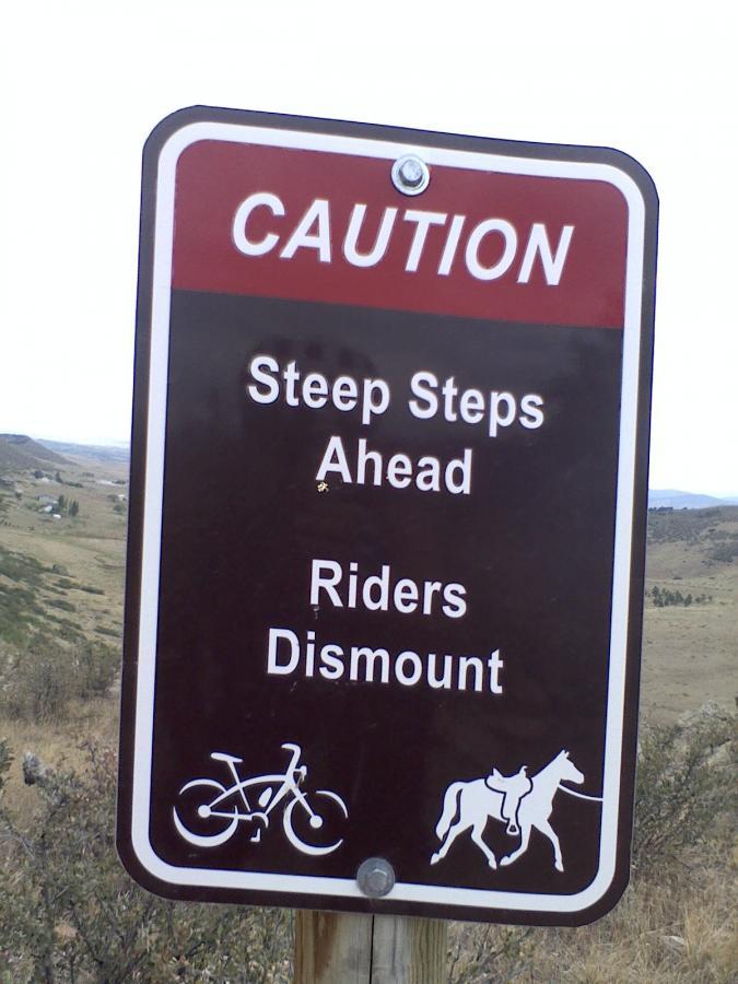 A caution sign indicating steep steps ahead, advising riders on bicycles and horses to dismount. The sign features icons of a bicycle and a horse. The background displays a hilly landscape. Coyote Ridge mountain bike trail.