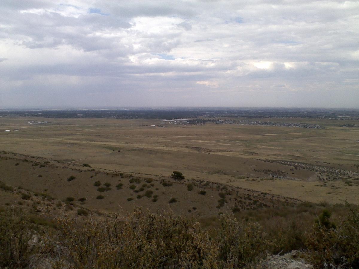 A panoramic view of an expansive, arid landscape under a cloudy sky. The foreground features rolling hills with sparse vegetation, leading into a broad, flat area in the distance that includes patches of grassland and scattered buildings. The horizon stretches far and wide, highlighting the vastness of the terrain. Coyote Ridge mountain bike trail.
