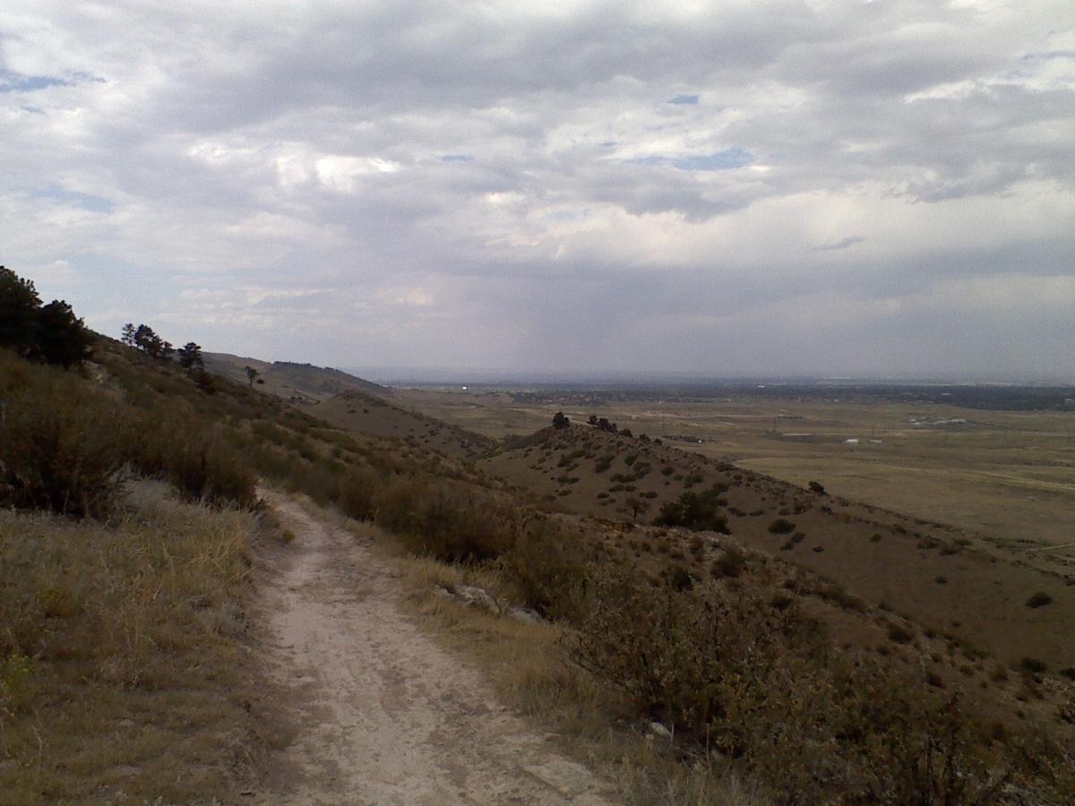 A scenic view of a dirt path winding through rolling hills, surrounded by sparse vegetation and low shrubs. The sky is overcast with clouds, and in the distance, a flat valley is visible, leading to a horizon lined with faint outlines of buildings. Coyote Ridge mountain bike trail.