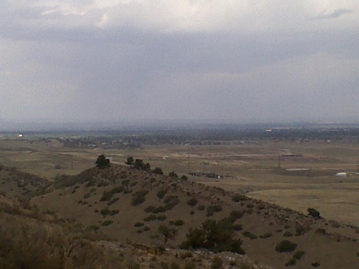 A panoramic view of rolling hills and a vast, open landscape under a cloudy sky. The foreground features gentle slopes with sparse vegetation, while the expansive background reveals distant hills and a hint of urban development below. Coyote Ridge mountain bike trail.