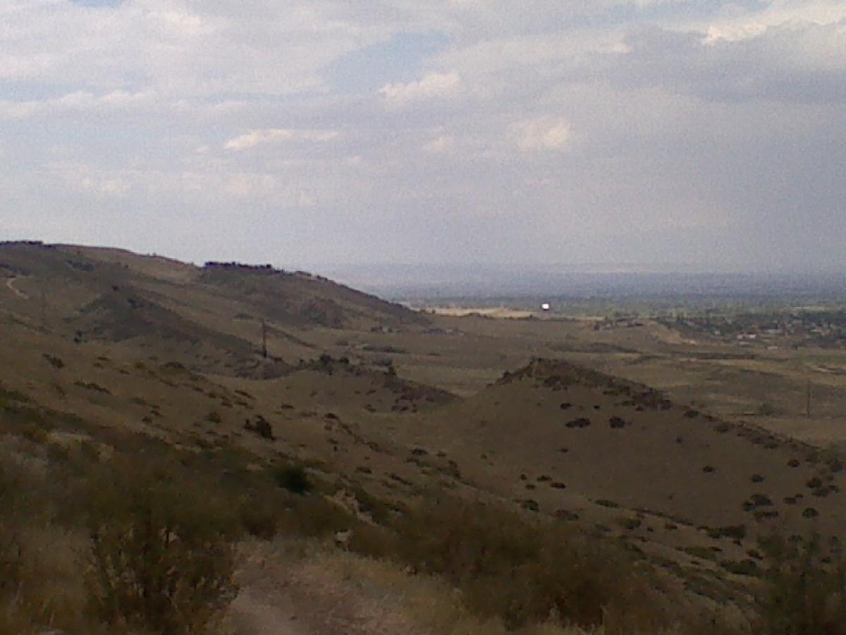 A panoramic view of rolling hills under a partly cloudy sky, showcasing a landscape with gentle slopes and sparse vegetation. In the distance, fields and a hint of a town can be seen, with power lines stretching across the terrain. The image captures the natural beauty and serenity of the countryside. Coyote Ridge mountain bike trail.
