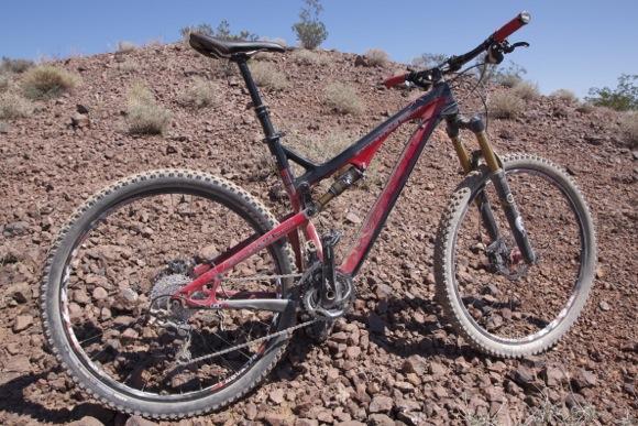 Intense Spider 29: A mountain bike resting on rocky terrain. The bike features a red and black frame, front suspension, and thick tires designed for off-road cycling. Sparse vegetation and blue sky are visible in the background.