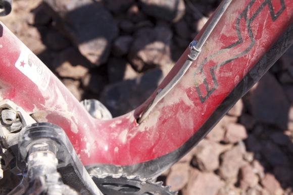 Intense Spider 29: Close-up view of a dusty red bicycle frame, highlighting the area where the frame connects to the bottom bracket. The surrounding rocky terrain is visible in the background.