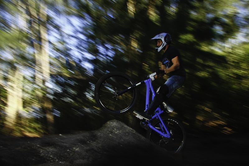A mountain biker performing a trick, with the front wheel lifted off the ground while riding a purple bike on a dirt ramp. The background features a blurred forest, creating a sense of motion and speed. Markham Park mountain bike trail.