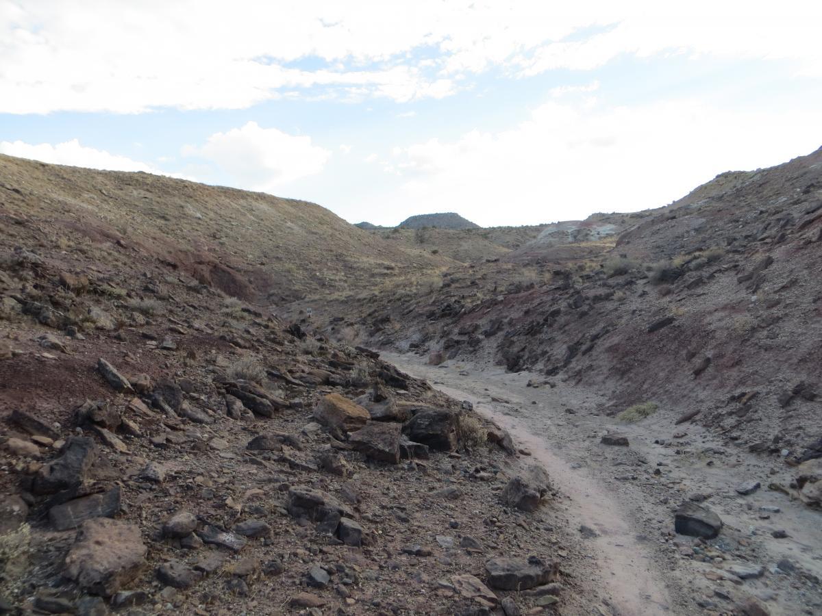 A rocky, uneven path winds through a dry, rugged landscape with hills and scattered boulders under a partly cloudy sky. The terrain appears arid, with sparse vegetation and earthy tones dominating the scene. Pet - A - Kes mountain bike trail.