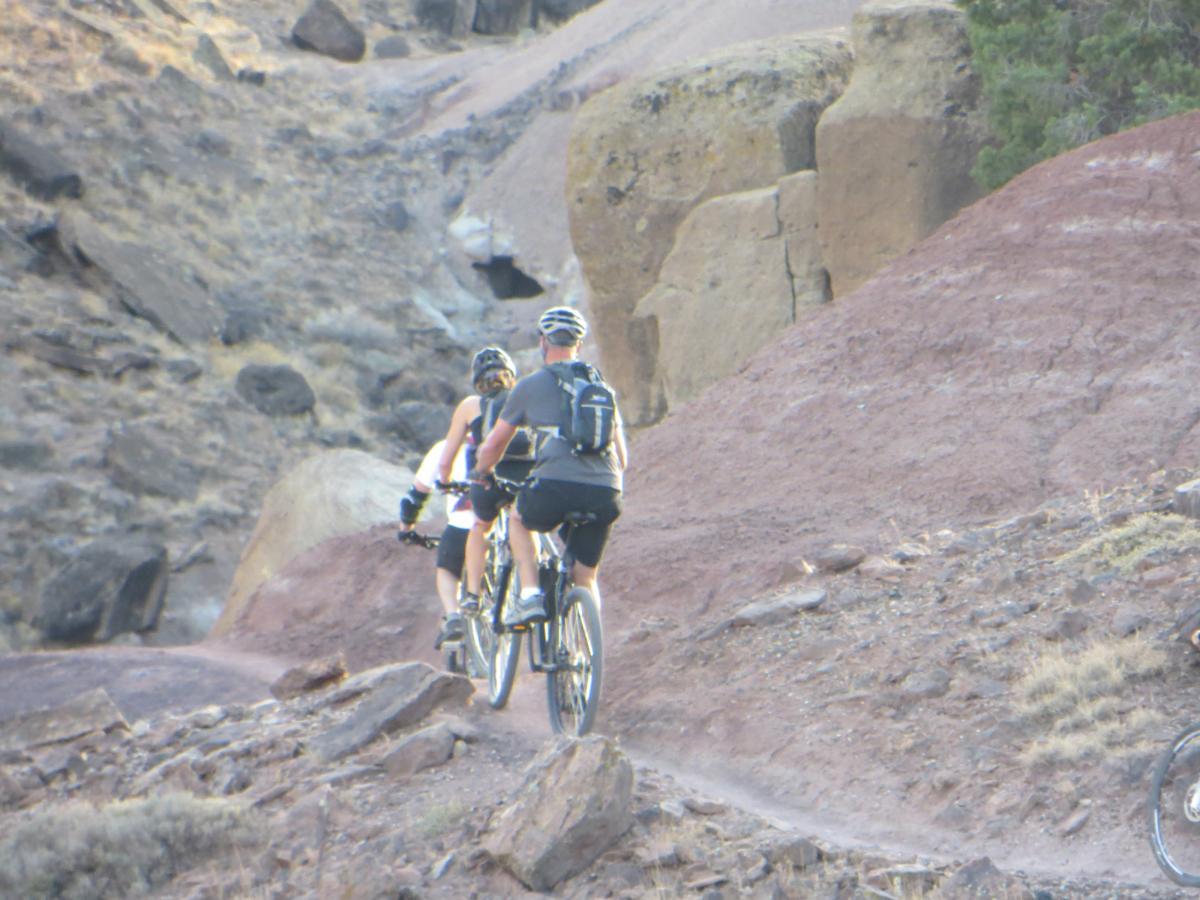Two mountain bikers ride along a rocky trail in a desert landscape, surrounded by earthy tones and steep terrain. The path winds through the rugged terrain, with patches of shrubs and large rocks in the background. Pet - A - Kes mountain bike trail.