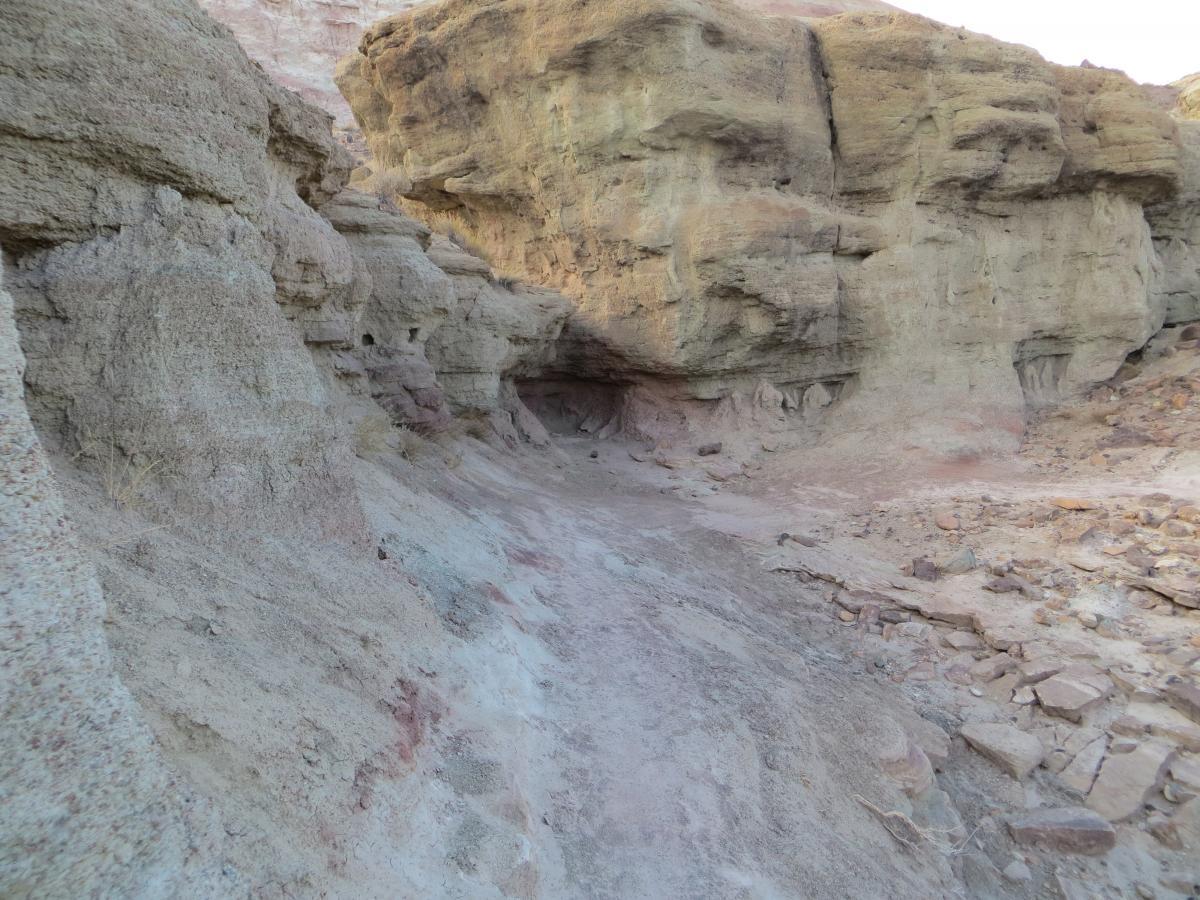 A rocky canyon landscape featuring layered, textured rock formations in shades of beige and brown, with a narrow path running through the center and surrounding cliffs. Pet - A - Kes mountain bike trail.