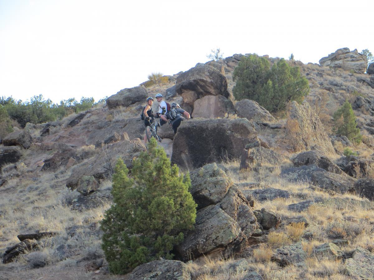 Two mountain bikers, wearing helmets and cycling gear, are posing on a rocky hillside with scattered shrubs and dry grass. They are surrounded by a rugged landscape featuring large boulders and sparse vegetation. The scene is set in a natural outdoor environment, indicating an adventurous mountain biking location. Pet - A - Kes mountain bike trail.