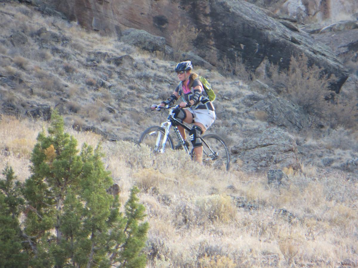 A person riding a mountain bike uphill on rocky terrain, surrounded by dry grass and shrubs. The rider is wearing a helmet and protective gear, with a backpack visible on their back. Pet - A - Kes mountain bike trail.
