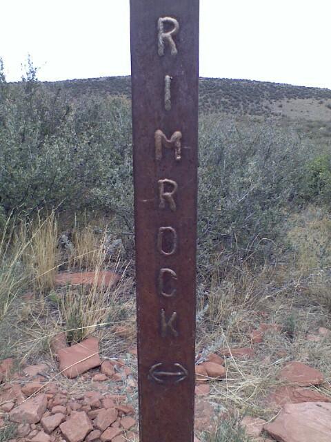 Rusty trail marker post with the word "RIMROCK" engraved vertically, set against a backdrop of sparse vegetation and rolling hills. Rimrock mountain bike trail.