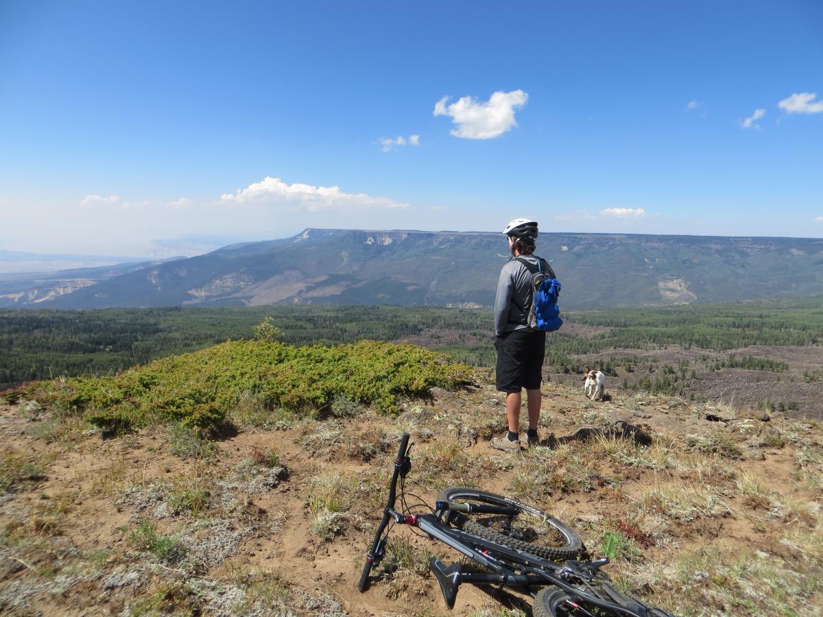 A person in a helmet and hiking attire stands on a rocky overlook, gazing at a vast mountain landscape. A mountain bike lies on the ground beside them, and a dog is visible in the background. The scene features a clear blue sky with a few clouds and lush green forests in the valley below. West Bench Trail mountain bike trail.